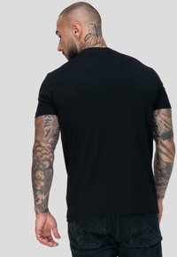 Mies, jolla on lyhyet hiukset ja tatuoinnit kaulassa ja käsivarsissa, pukeutuneena yksinkertaiseen mustaan t-paitaan ja tummaan farkkuihin, kääntynyt pois vaaleanharmaata taustaa vasten.