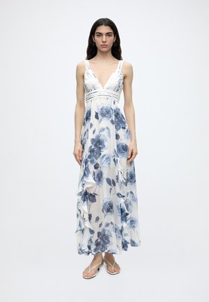 Γυναίκα με μακριά σκούρα μαλλιά φοράει λευκό φόρεμα μίντι με μπλε λουλουδάτα σχέδια, κροσέ μπούστο και φούστα με βολάν, συνδυασμένο με λευκά σανδάλια.