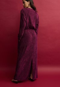 Robe maxi couleur prune avec une texture à côtes verticales, manches longues, fente sur le côté et taille élastique. Portée avec des sandales à talons.