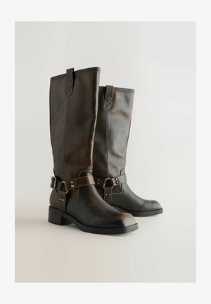 Brune læderknæhøje støvler med en glat finish, spændeaccent og dekorativ syning i siderne. Blokhælen giver stabilitet.