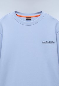 Lyseblå genser med ribbestrikket rund hals, oransje innvendig fôr, brodert logo på brystet, og myk tekstur.