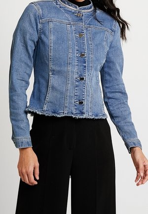 Kvinde iført en tætsiddende blå denimjakke med metalknaplukninger og frynset kant, kombineret med sorte bukser med vide ben.