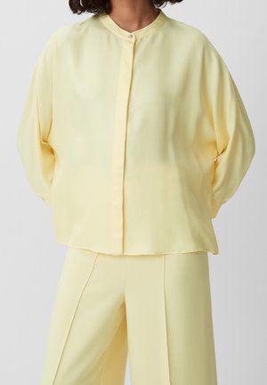 Mujer que lleva una blusa suelta de manga larga de color amarillo pálido con botones delanteros y pantalones anchos a juego sobre fondo gris.