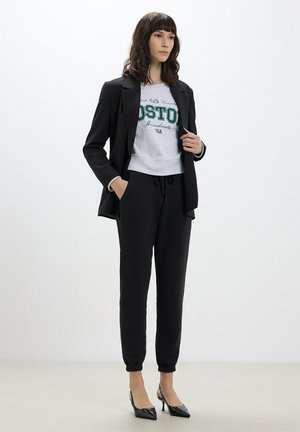 Blazer nero su una felpa grigia corta con stampa "BOSTON", abbinato a pantaloni neri stile jogger e tacchi a punta neri.