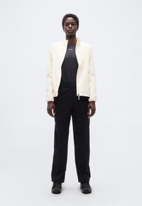 Άτομο όρθιο, ντυμένο με μπεζ ζακέτα με φερμουάρ, μαύρο μπλουζάκι Salomon, μαύρο παντελόνι και μαύρα παπούτσια πεζοπορίας, σε απλό λευκό φόντο.