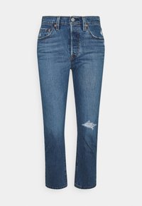 Jeans de mezclilla azul con un corte recto, que presentan un parche desgastado cerca de la rodilla y un diseño estándar de cinco bolsillos. Cierre clásico de botón y cremallera.