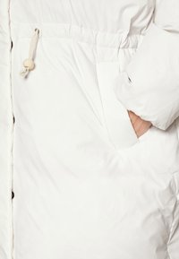 Λευκό χνουδωτό παλτό με απαλό υφή, διαθέτοντας ελαστική κορδέλα στη μέση και πλαϊνή τσέπη με καπάκι.