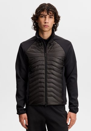 Jonge man met krullend haar, gekleed in een zwart jack met ritssluiting en doorgestikte voorpanelen, afgebeeld tegen een eenvoudige lichte achtergrond.