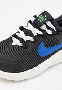 Fekete futócipő bordázott textúrával, fehér fűzőkkel és kék Nike logóval. Zöld csillag alakú logó található a nyelvén.