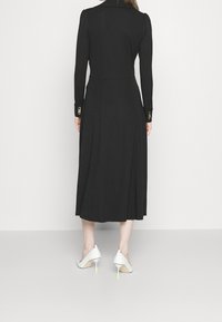 Robe noire à manches longues avec un corsage ajusté et une jupe évasée. Présente des accents de boutons dorés sur les poignets. Associée à des chaussures à talons blanches.