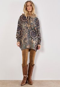 Túnica con capucha y de manga larga en tono beige que presenta intrincados patrones en azul y burdeos. Combinada con mallas de ante marrón y botas de vaquero.