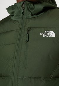 Tumma vihreä pehmustettu takki, jossa on vetoketjuita edessä, korkea kaulus ja brodeerattu logo rinnassa; siinä on teksturoitu kangas ja kestävät vetoketjut.