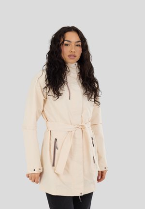 Donna con capelli lunghi e ricci che indossa un cappotto beige chiaro con cintura, tasche con cerniera e polsini con bottoni, in piedi davanti a uno sfondo uniforme.