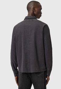 Uomo che indossa una maglietta a maniche lunghe grigia scura e jeans neri, di spalle, in piedi contro uno sfondo grigio chiaro uniforme.
