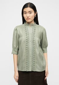 Γυναίκα που φοράει ανοιχτό πράσινο πουκάμισο με κουμπιά και δαντέλα στο τελείωμα, μανίκια 3/4, σε συνδυασμό με σκούρα φούστα, σε λευκό απλό φόντο.