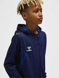 Jongen met blonde dreadlocks die een donkerblauwe Hummel-hoodie met ritssluiting en logo draagt, kijkt naar rechts tegen een effen lichte achtergrond.