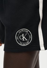 Gros plan sur une personne portant un short noir texturé avec une bande blanche et un patch rond avec le logo Calvin Klein près de l’ourlet.