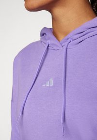 Fialová mikina vyrobená z bavlneného zmesi s kapucňou na šnúrku a malým strieborným logom Adidas na hrudi. Hladká textúra.