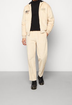 Beige corduroy jas met zwarte accenten en borduurwerk, gecombineerd met bijpassende beige corduroy broek en zwarte veterschoenen.