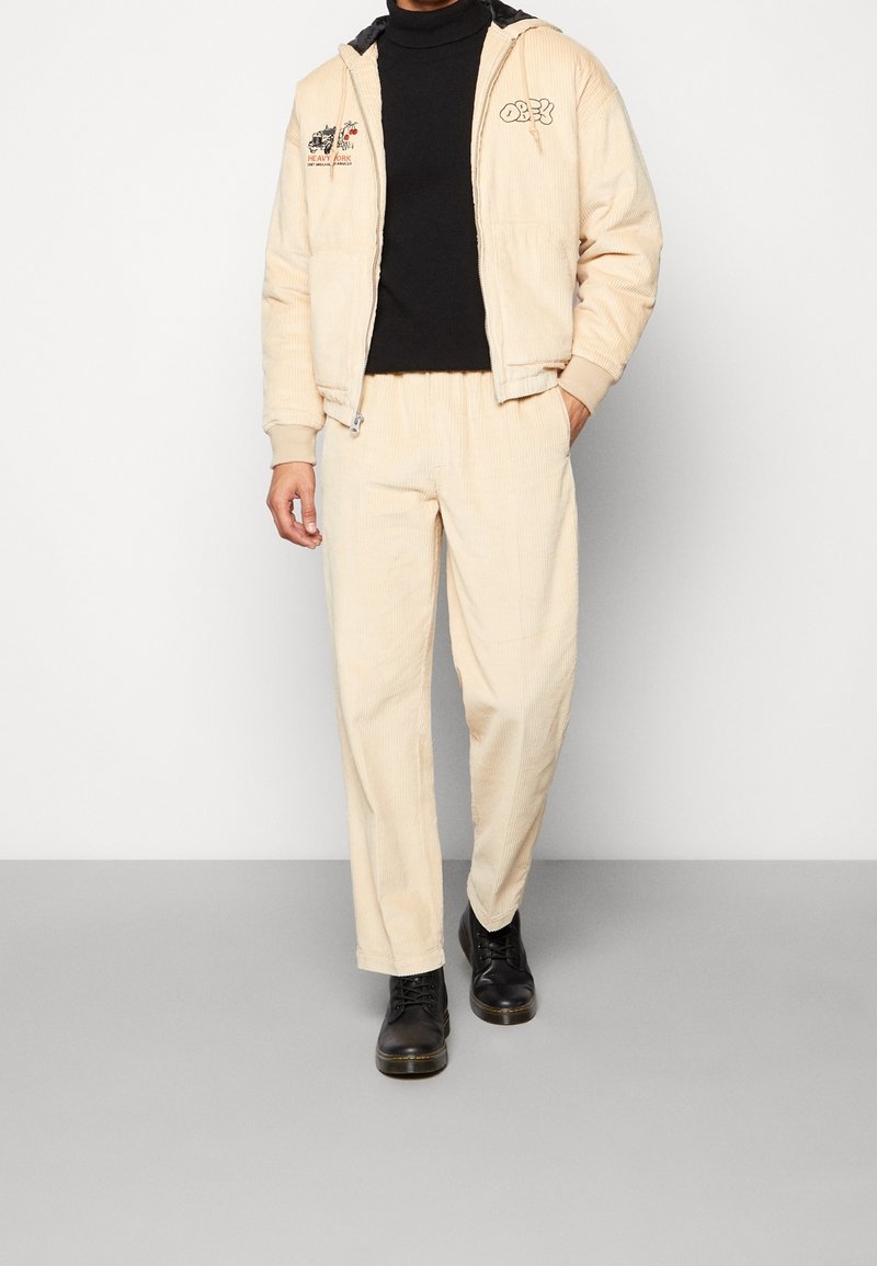 Beige kordflöjlsjacka med svarta accenter och broderier, matchad med beige kordflöjlsbyxor och svarta snörboots.