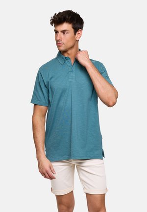 Hombre ajustando el cuello de una camiseta polo de manga corta color verde azulado, llevando pantalones cortos beige remangados, de pie contra un fondo liso y claro.