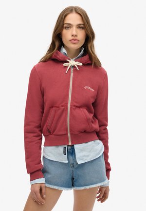 Jeune femme avec des cheveux bruns lâches portant un sweat à capuche rouge zippé sur une chemise à rayures bleues et un short en denim clair sur un fond uni.