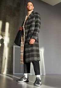 Plaid overcoat in black and gray with white accents, layered over a tan turtleneck. Paired with black athletic shoes and cropped pants.