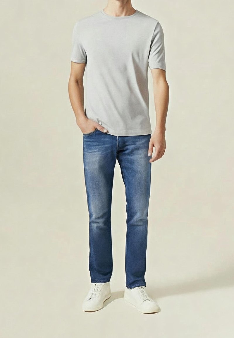 Homme portant un t-shirt gris clair à manches courtes, jean bleu et baskets blanches, debout avec une main dans la poche.