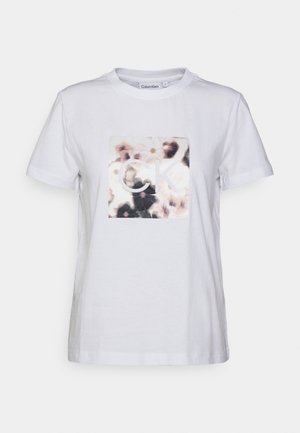 Camiseta blanca de algodón con un diseño floral que incluye un gráfico cuadrado y las letras "CK" en el centro. Manga corta y cuello redondo.
