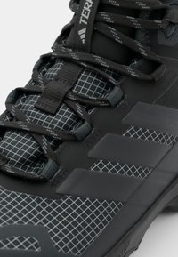Schwarzer Sportschuh mit strukturiertem Mesh-Obermaterial, Gittermuster, flachen Schnürsenkeln und drei markanten dunklen Streifen an der Seite.