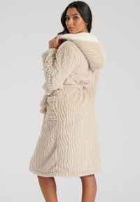 Beige, gehooded badjas van gestructureerde, pluizige stof. Heeft een ceintuur in de taille en zijzakken, met een zachte, contrasterende binnenvoering.