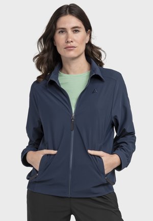 Frau mit schulterlangem braunem Haar, die eine marineblaue Jacke mit Reißverschluss über einem hellgrünen Hemd trägt, die Hände in den Jackentaschen, neutraler Hintergrund.