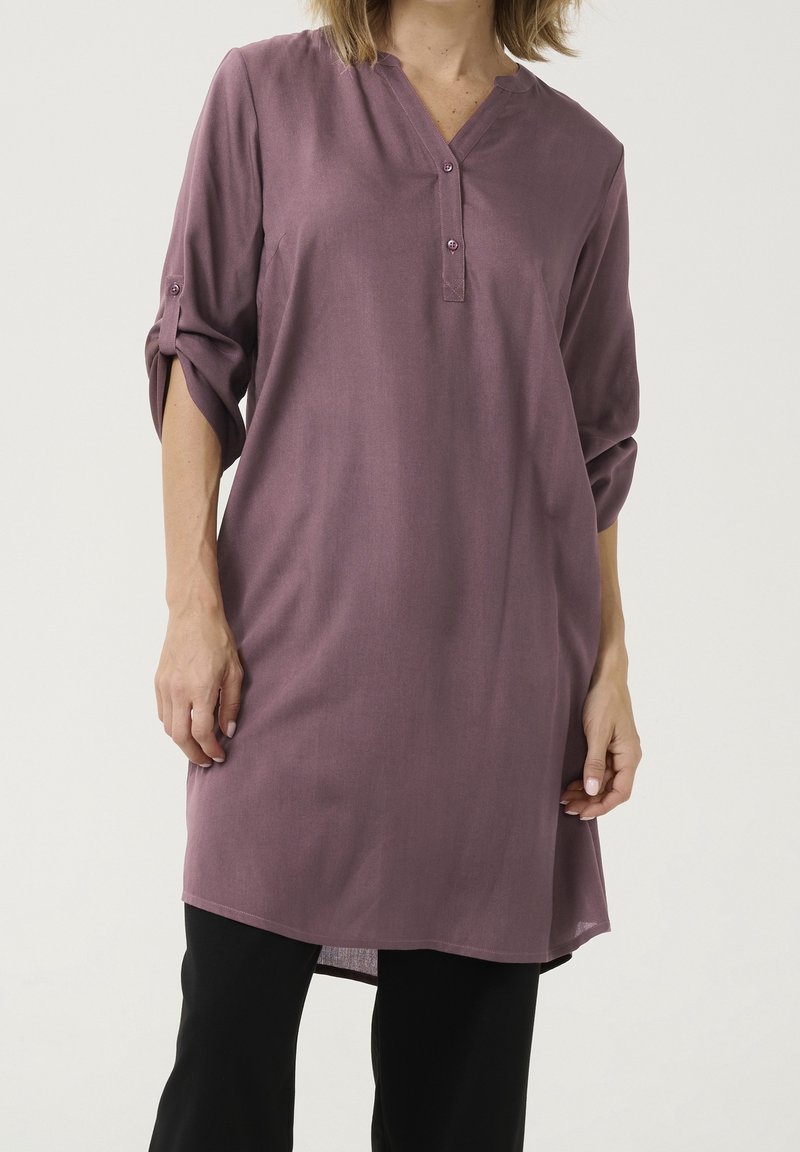 Femme portant une tunique mauve ample à manches longues avec les manches retroussées et un pantalon noir sur un fond clair uni.