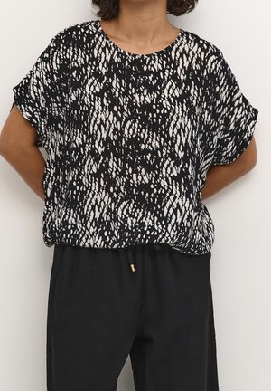 Femme portant un chemisier à manches courtes à motif noir et blanc et un pantalon noir ample, debout les mains dans le dos.