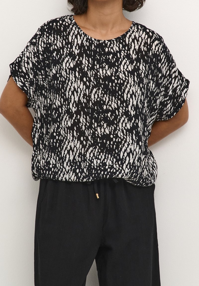 Femme portant un chemisier à manches courtes à motif noir et blanc et un pantalon noir ample, debout les mains dans le dos.