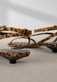 Bruna sandaler med leopardtryck som har en mjuk pälsstruktur, svarta remmar och små, runda klackar. Har design med öppen tå och tunna vristremmar.