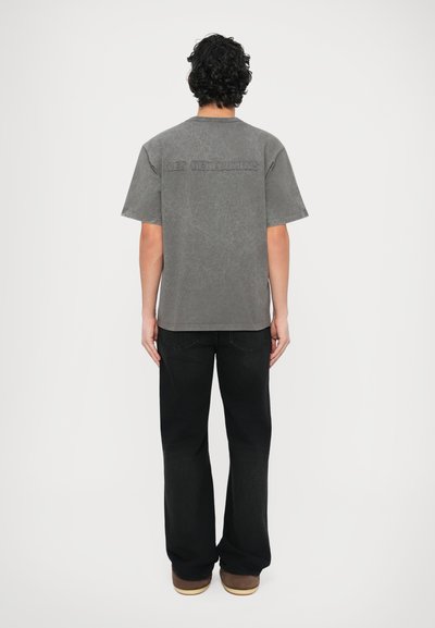 Szürke, rövid ujjú póló laza szabással és dombornyomott szöveggel a hátulján. Széles szárú fekete farmerrel és barna cipővel párosítva.