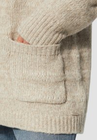 Beige gestrickte Strickjacke mit gerippten Bündchen und einem strukturierten Oberteil, mit einer Fronttasche und einem metallischen Logo-Akzent nahe dem Saum.