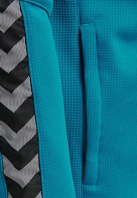 Teal træningsjakke med tekstureret stof, ribbet mønster og en sort sidepanel med hvid stribet zigzagdesign.