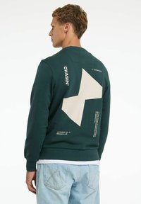 Felpa verde realizzata in tessuto testurizzato, con un grande motivo geometrico beige e testo stampato sul retro. Indossata con jeans azzurri chiari.