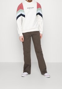 Valkoinen collegepaita, jossa on punaisia, vihreitä ja tummansinisiä raitoja sekä painettu logo. Yhdistetty ruskeisiin housuihin ja valkoisiin tennareihin.