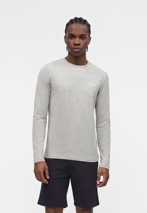 Bărbat tânăr cu părul împletit, purtând o cămașă gri deschis cu mâneci lungi și pantaloni scurți negri, stând încrezător în fața unui fundal simplu.