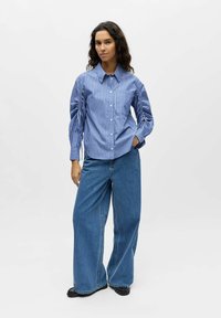 Chemise à rayures bleues et blanches avec manches bouffantes, associée à un jean en denim ample et des sandales noires. Tenue simple et décontractée.
