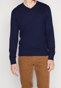 Marinblå V-ringad tröja i mjukt stickat material, med långa ärmar och ribbad nederkant, tillsammans med bruna manchesterbyxor.