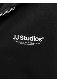 Crni dukserica s bijelim ispisanim tekstom "JJ Studios" i dodatnim tekstom ispod. Tkanina je glatka s detaljom na rajsferšlusu.