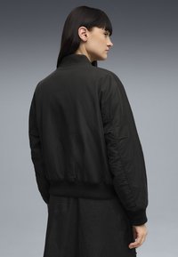 Veste bomber noire avec col et poignets côtelés, en matériau matelassé, et une poche latérale. Texture lisse, coupe décontractée et design raccourci.