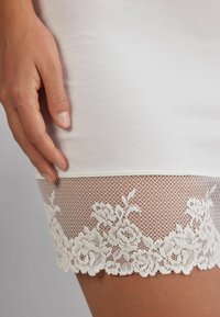 Vit klänning med spetsfåll med blommönster och ett texturerat mesh-design. Släta tyg med en elegant, kroppsnära passform.