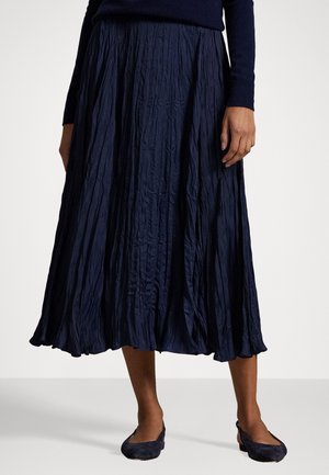 Mujer vestida con una falda larga plisada azul marino, top de manga larga a juego y zapatos azul marino de tacón bajo, de pie sobre un suelo claro.