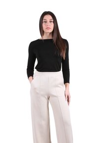 Donna con capelli lunghi e lisci che indossa un top nero aderente a maniche lunghe e pantaloni beige a gamba larga con le mani nelle tasche su sfondo bianco.