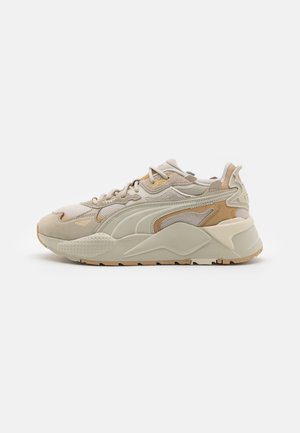 Scarpa da ginnastica beige e marrone chiaro con dettagli in mesh e camoscio, lacci bianchi, suola spessa imbottita e battistrada testurizzato, vista laterale su sfondo bianco.